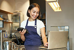 Eine junge Frau sitzt in einem Café an einem Laptop und hat ein mobiles Bezahlterminal in der Hand.