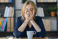 Eine gestresste Frau reibt sich am Arbeitsplatz müde die Augen (Symbolbild).