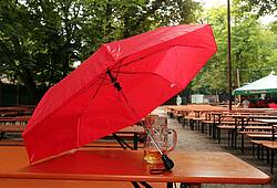 Ein Maßkrug steht in einem Biergarten bei Regenwetter unter einem roten Regenschirm.