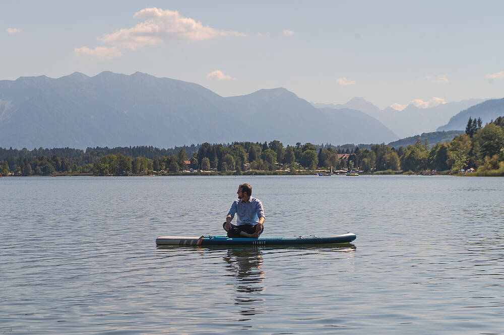 Ein Stand-up-Paddler sitzt auf einem oberbayerischen See auf seinem Board, im Hintergrund die Berge.