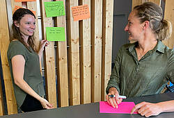 Zwei Frauen unterhalten sich vor einer Pinnwand mit Post-its.