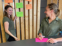 Zwei Frauen unterhalten sich vor einer Pinnwand mit Post-its.