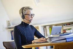 Eine Frau mit Headset sitzt an einem Schreibtisch vor ihrem Laptop (Symbolbild).