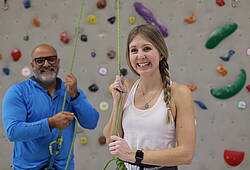 Eine junge Frau und ein älterer Mann stehen vor einer Kletterwand. Der Mann sichert die Frau mit einem Seil.