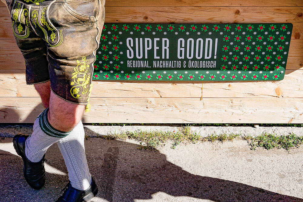 Ein Mann in Lederhose lehnt auf dem Münchner Oktoberfest 2023 an einem Wiesn-Standl. Zu sehen sind nur die Beine und ein am Standl angebrachtes Schild mit der Aufschrift „Super Good! Regional, nachhaltig & ökologisch“. Das Standl ist vom Münchner Bauern Stadl, dort gibt es Semmeln mit Quinoa von der Münchner Bauern Genossenschaft.