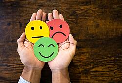 Zwei Hände halten drei Smileys aus Karton mit einem lachenden, neutralen und traurigen Gesicht (Symbolbild).