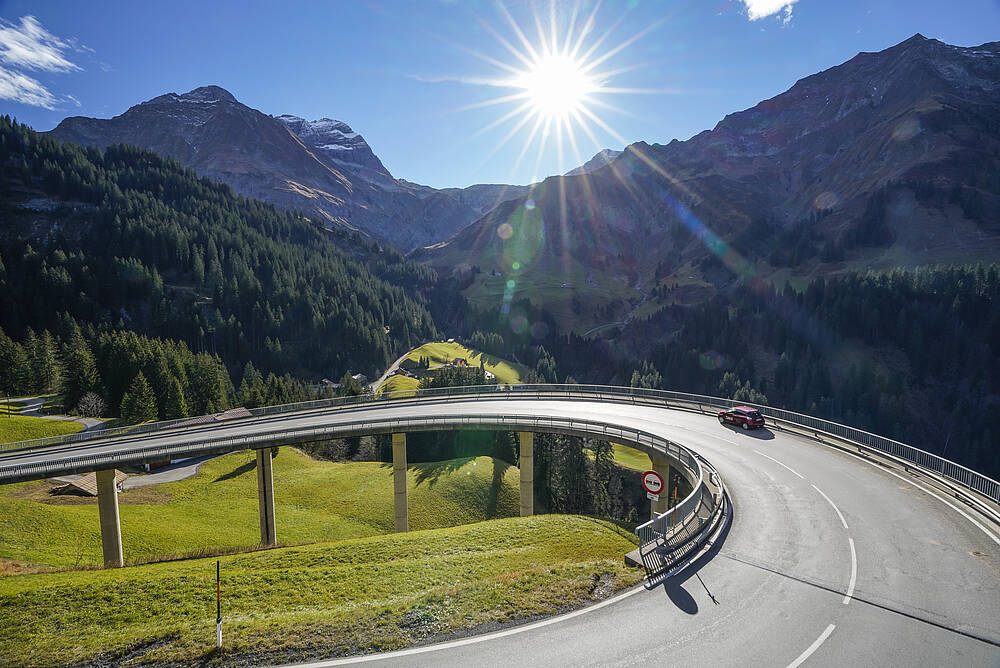 Ein Auto fährt auf einer Alpenstraße bei Sonnenschein durch die Berge.