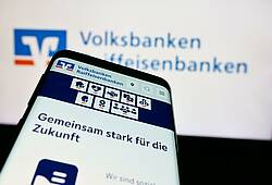Symbolfoto für die VR Banking App: Ein Smartphone mit der VR Banking App und dem Schriftzug "Gemeinsam stark für die Zukunft" wird vor einen Bildschirm mit dem Schriftzug der Volksbanken Raiffeisenbanken gehalten.