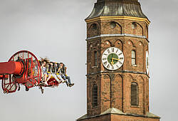 Oktoberfestbesucher in einem Fahrgeschäft fliegen durch die Lüfte, im Hintergrund ein Münchner Kirchturm.