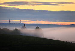 Zwei Windräder und eine Kirche versinken im Nebel (Symbolbild für Energiewende).